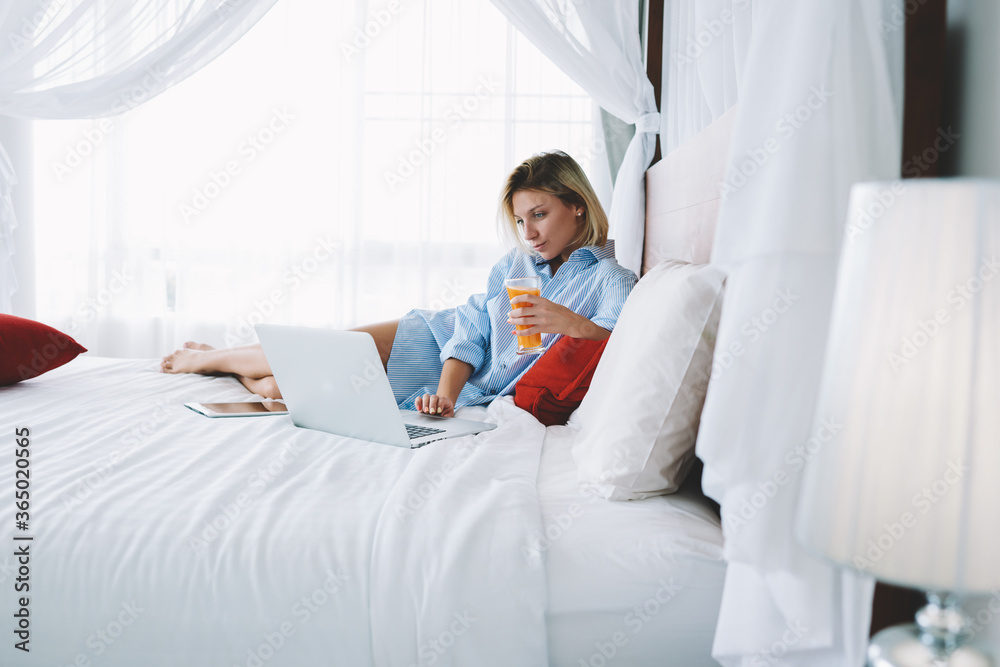 Young woman watching movie on laptop computer relaxing in bed with fresh juice in morning, female freelancer enjoying remote work using netbook connected to wifi for read news recreating in hotel room Young woman watching movie on laptop computer relaxing in bed with fresh juice in morning, female freelancer enjoying remote work using netbook connected to wifi for read news recreating in hotel room