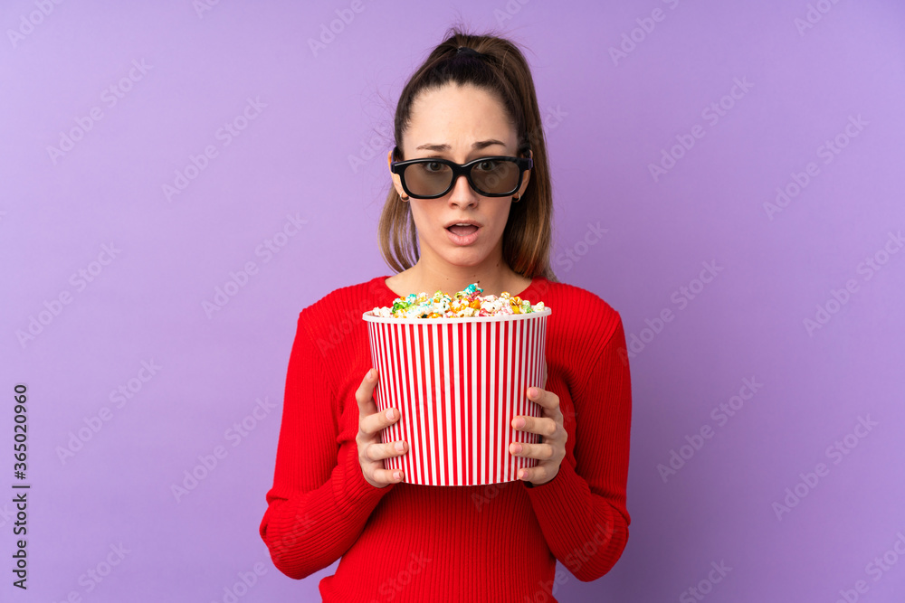 Young brunette woman over isolated purple background surprised with 3d glasses and holding a big bucket of popcorns Young brunette woman over isolated purple background surprised with 3d glasses and holding a big bucket of popcorns