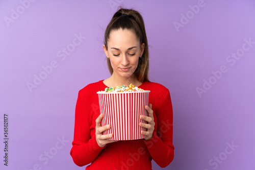 Young brunette woman over isolated purple background holding a big bucket of popcorns