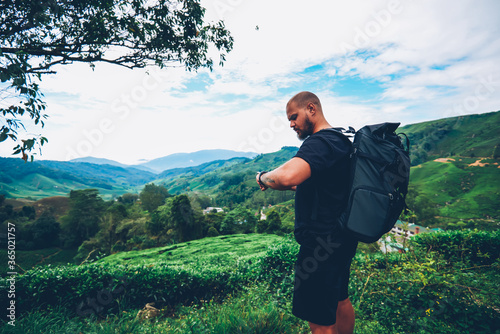 Billede på lærred Pensive bearded tourist with backpack looking at smartwatch to tracking GPS walking in green locality of asia mountains