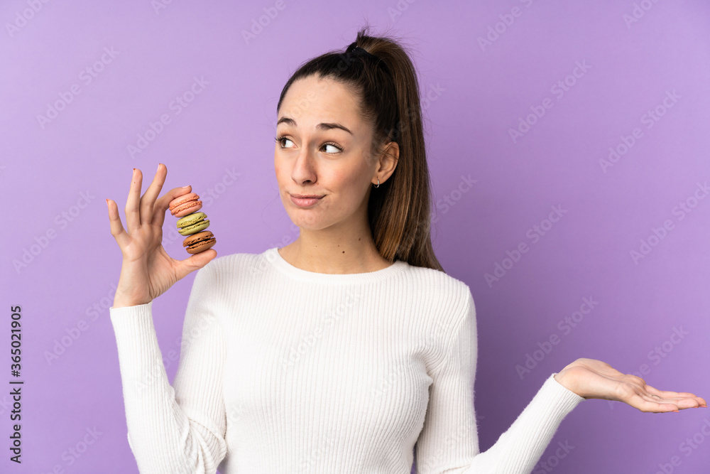 Young brunette woman over isolated purple background holding colorful French macarons and making doubts gesture Young brunette woman over isolated purple background holding colorful French macarons and making doubts gesture