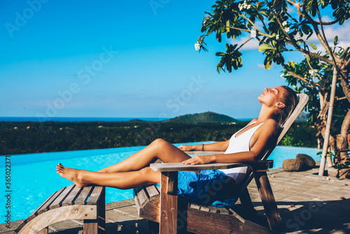 Attractive slim young woman dressed in stylish white swimsuit sunbathing on wooden sunbed near blue swimming pool enjoying summer resort.Female sexy tourist tans near basin thanks to hot trip photo