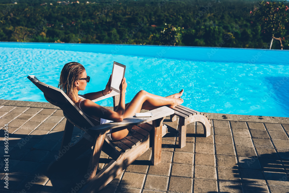 Young attractive woman in sunglasses reading news on website on modern touch pad device with blank screen area connected to 4G internet while resting on sunbed and enjoying summertime during vacation