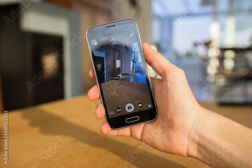 A hand holding a mobile phone which displays the camera app on the touch screen. The image is taken in the living room of a home.