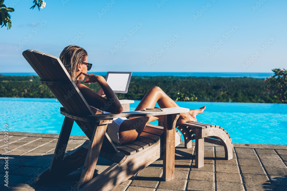 Pensive young woman in sunglasses reading email and working remotely using modern tablet with blank screen area for your advertising content while relaxing on sunbed after swimming in blue pool