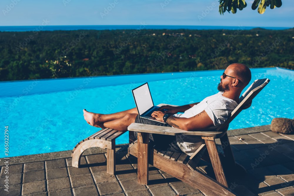 Bearded businessman lying near blue pool while typing text on keyboard of netbook with blank copy space screen for advertising content working remotely during summer resort in tropical locality