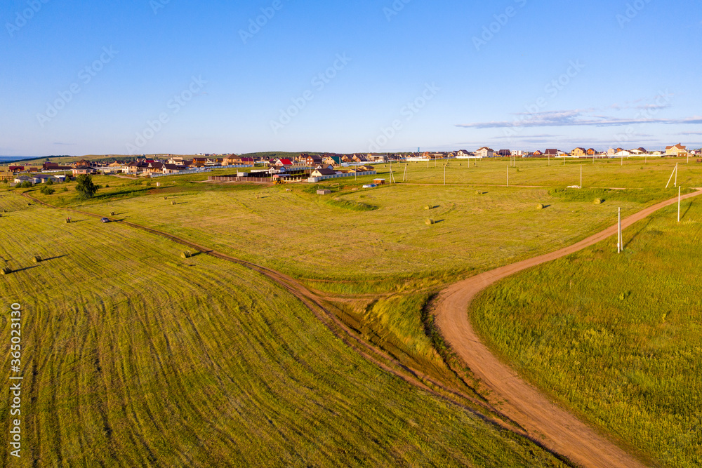 Rural landscape. In the foreground is a meadow with bales of mown grass, in the background is a rural settlement with a power line. Shooting from a drone.