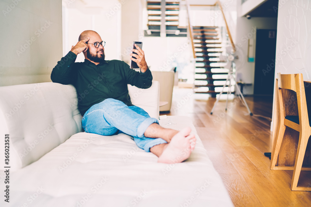 Young hipster guy in casual wear lying on cozy sofa reading interesting book online resting at modern interior apartments, pensive bearded male enjoying watching movie on portable pc at home