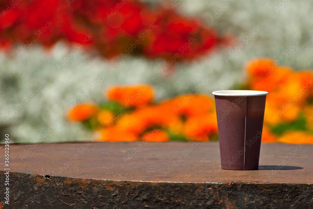 Brown paper cup on a background of greenery. Coffee on a marble border. Flowers and flower beds of the city are blurred in the background. Sharpness on coffee. Layout for design.