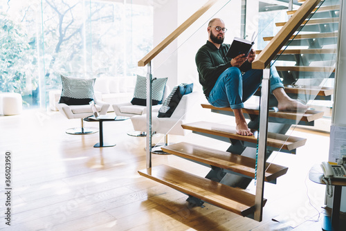 Young bearded guy fond of literature resting with favorite book at coy home interior, bearded man in spectacles reading interesting bestseller sitting at stairs at modern designed apartments