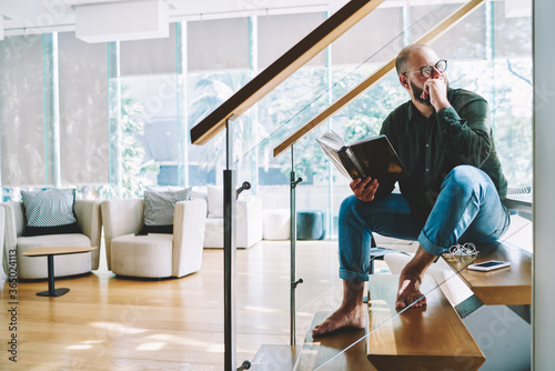 Pensive male in eyewear analyzing novel after finishing reading novel spending free time at home on weekends, thoughtful hipster guy fond of literature sitting at stairs at modern designed apartment