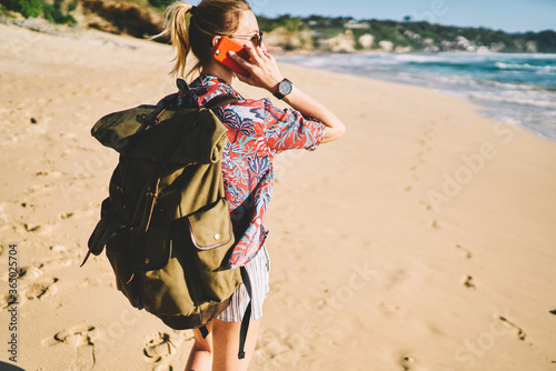 Back view image of young female tourist dressed in casual clothes communicating with friends via application and roaming on smartphone.Woman traveler enjoy vacation while walking on sandy beach