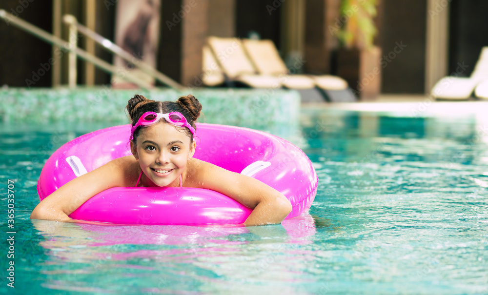 Happy little girl in goggles playing with a pink inflatable ring in the swimming pool on a hot summer day. Kids learn to swim. Child water toys.Family beach vacation.