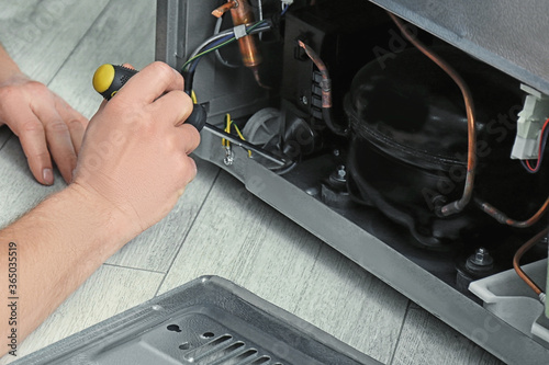 Repairman with screwdriver fixing refrigerator indoors, closeup photo