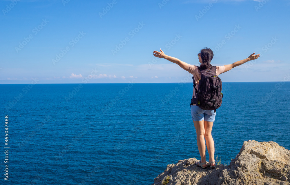 brunette woman in t-shirt with backpack stands back on rocky seaside arms outstretched, and looks out at the sea. Solo summer outdoor activities in fresh air. Concept of Hiking trekking. Copy space brunette woman in t-shirt with backpack stands back on rocky seaside arms outstretched, and looks out at the sea. Solo summer outdoor activities in fresh air. Concept of Hiking trekking. Copy space