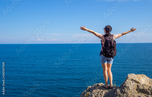 brunette woman in t-shirt with backpack stands back on rocky seaside arms outstretched, and looks out at the sea. Solo summer outdoor activities in fresh air. Concept of Hiking trekking. Copy space © Tani_Bel