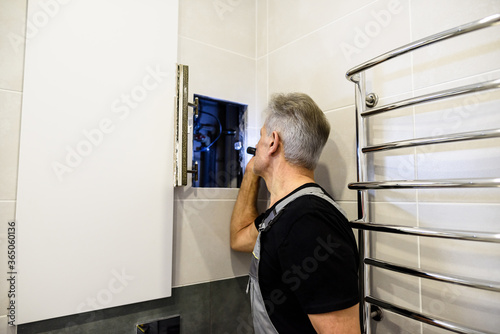 Slika na platnu Aged plumber, repairman in uniform examining, inspecting pipes using flashlight in the bathroom