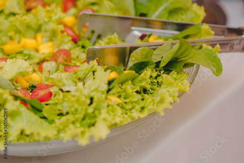 salada de folhas de alface com tomate e milho em refrat  rio de vidro em cima de uma mesa branca e um pegador de inox apoiado 