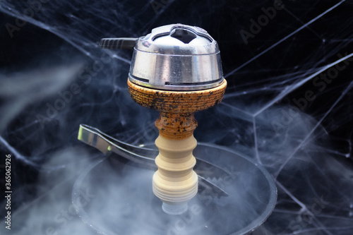 Bowl of hookah with kaloud lotus, saucer and tongs, smoke around, black background with spider web.