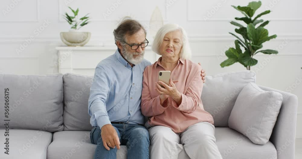 Old Caucasian cheerful couple sitting on couch and watching something on smartphone screen and talking. At home. Senior retired man and woman using mobile phone, tapping and scrolling, discussing. Old Caucasian cheerful couple sitting on couch and watching something on smartphone screen and talking. At home. Senior retired man and woman using mobile phone, tapping and scrolling, discussing.
