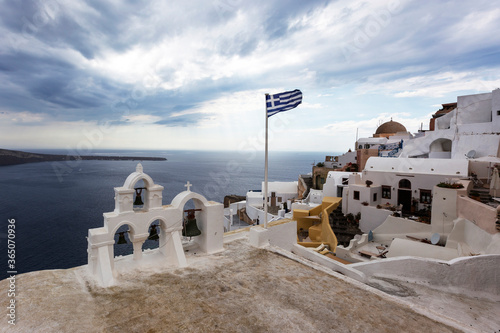 Santorini - Oia, view from the cliff to the caldera. White bell tower on the right and a white roof on the left on the cliff of Greek white houses. In the background an island and dramatic clouds.