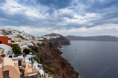 Santorini - Oia, view from the cliff to the caldera. On the left, a stone path that leads to white Greek houses that stand on a cliff. In the background the sea and the island.