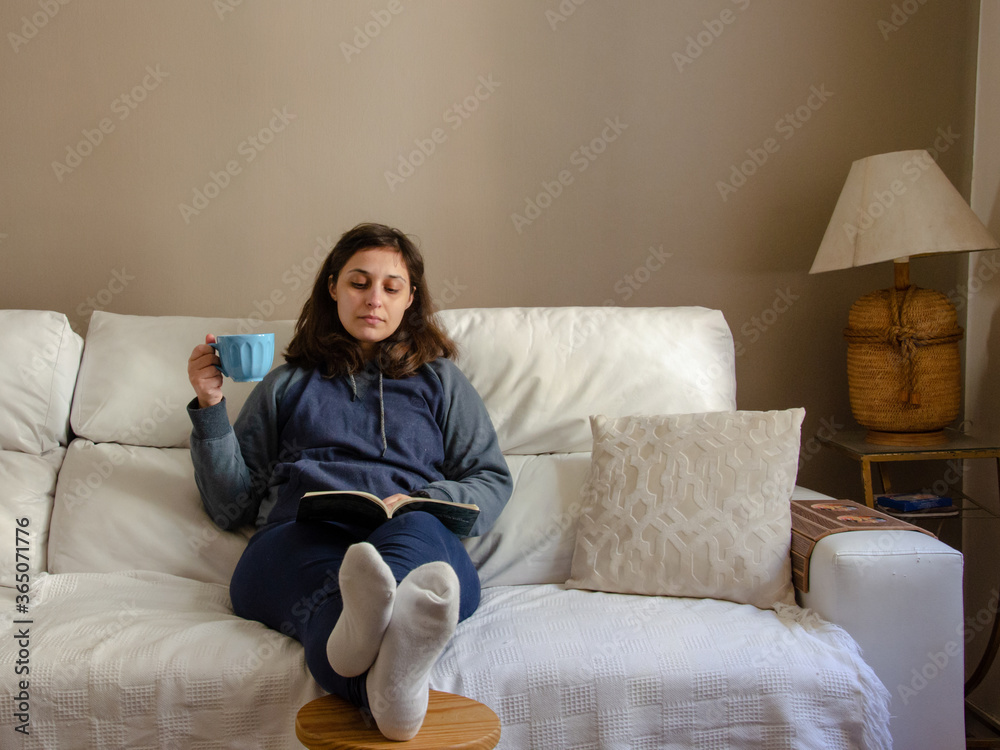 young woman sitting on a white sofa reading a book holding a coffee mug using comfort clothes young woman sitting on a white sofa reading a book holding a coffee mug using comfort clothes