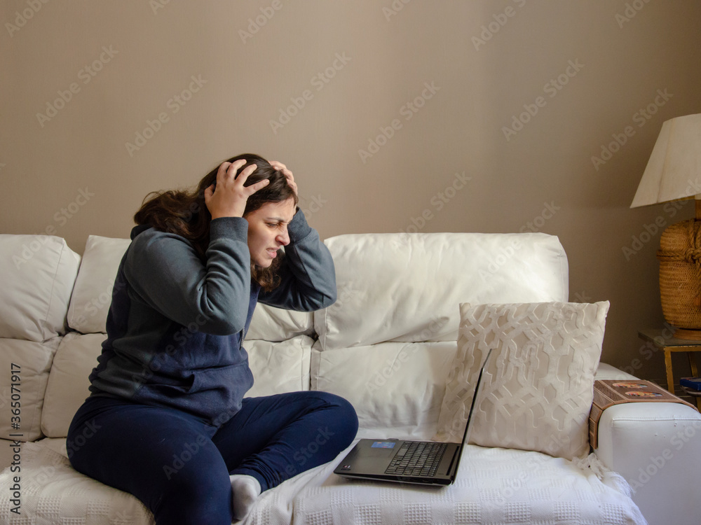 young woman sitting on a white sofa working on a laptop using comfort clothes looking worried with hands in head young woman sitting on a white sofa working on a laptop using comfort clothes looking worried with hands in head