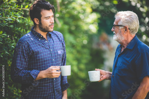 An adult hipster son and senior father talking at home, morning nature scene in the home garden outdoor