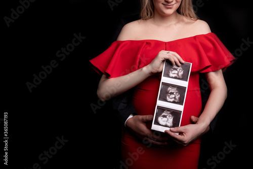 Pregnant woman in red dress holding ultrasound image. Concept of pregnancy, health care, gynecology, medicine. Young mother waiting of the baby. Close-up, copy space, indoors.