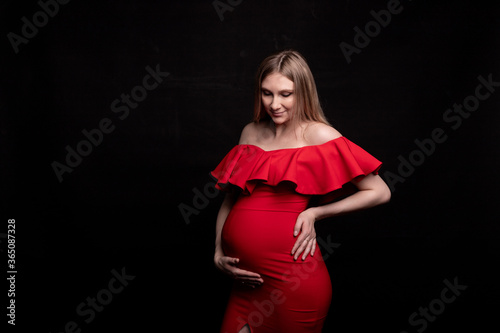 Pregnant woman in red dress holds hands on belly on a black background. Pregnancy, maternity, preparation and expectation concept. Close-up, copy space. Beautiful tender mood photo of pregnancy.