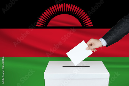 Election in Malawi. The hand of man putting his vote in the ballot box. Waved Malawi flag on background. 