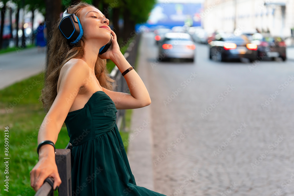 Young beautiful girl is listening to music on headphones. Against the background of a metal fence and road
