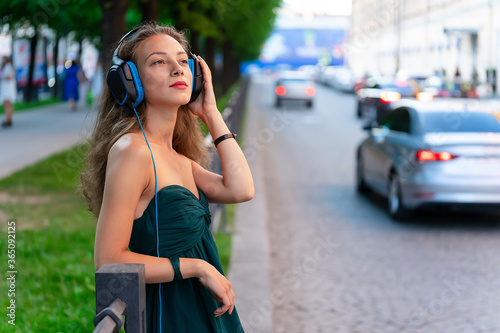 Young beautiful girl is listening to music on headphones. Against the background of a metal fence and road