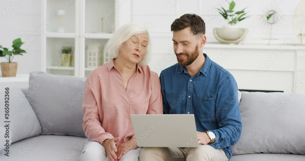 Caucasian young handsome adult son teaching his old gray-haired mother to use laptop on couch in cozy living room. Senior woman watching man using computer, explaining and typing. Indoors. Caucasian young handsome adult son teaching his old gray-haired mother to use laptop on couch in cozy living room. Senior woman watching man using computer, explaining and typing. Indoors.