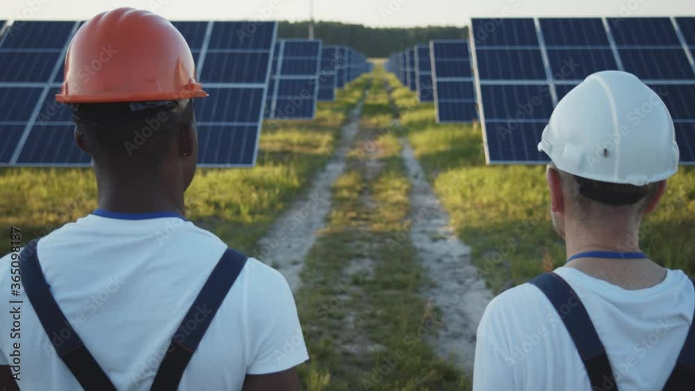 Caucasian and afro-american electrician workers walk between solar panels and discussing their work. New ecological construction. Cooperation. Team. Caucasian and afro-american electrician workers walk between solar panels and discussing their work. New ecological construction. Cooperation. Team.