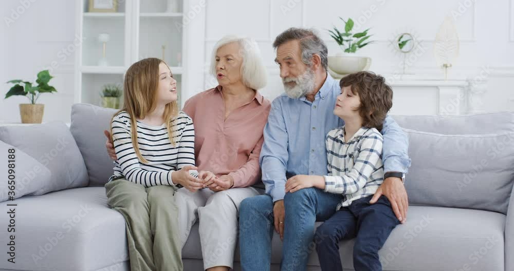 Caucasian old grandparents with grandchildren sitting on couch in cozy living room. Grandmother, grandfather, grandson and granddaughter spending time together at home. Indoors. Family generations. Caucasian old grandparents with grandchildren sitting on couch in cozy living room. Grandmother, grandfather, grandson and granddaughter spending time together at home. Indoors. Family generations.