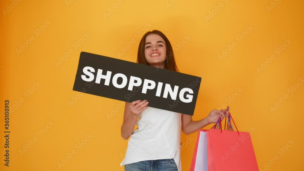 excited beautiful young woman with black shopping sign and colorful paper bags moving on yellow background