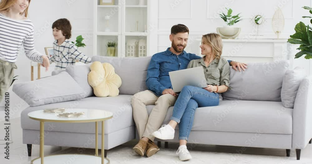 Two cheerful Caucasian kids running in circles and rounds in living room. Happy cute small sister and brother playing around sofa. Smiled mother and father sitting on couch with laptop computer. Two cheerful Caucasian kids running in circles and rounds in living room. Happy cute small sister and brother playing around sofa. Smiled mother and father sitting on couch with laptop computer.