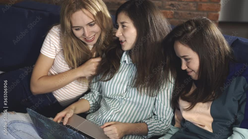 Multiracial people sitting on couch or sofa, looking at laptop screen and laughing. Charming women friends spend time at home at computer together. The concept spending time at home together Multiracial people sitting on couch or sofa, looking at laptop screen and laughing. Charming women friends spend time at home at computer together. The concept spending time at home together