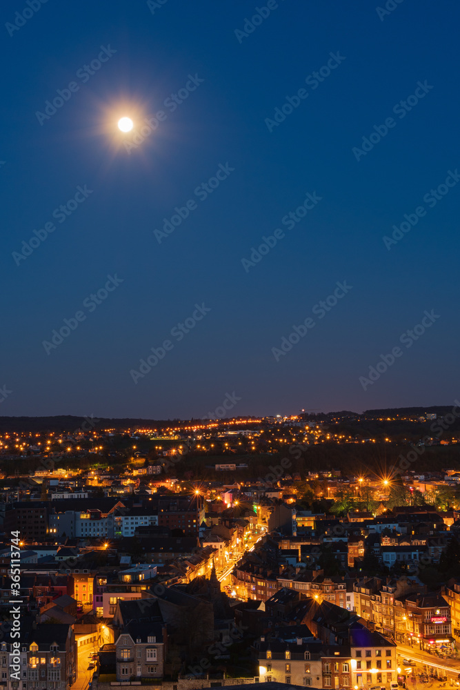 The moon shines upon Jambes as the sun sets and the city lights are switched on.