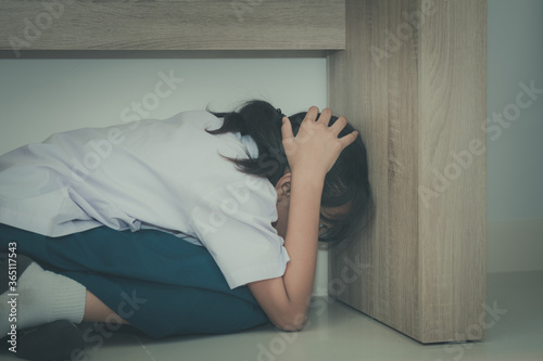 Asian young girl in a school uniform hides under the desk when an earthquake occurs with concept

