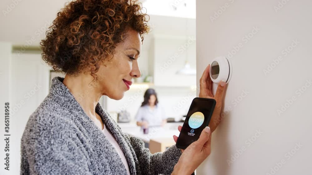 Mature woman using app on phone to control digital central heating thermostat at home with gay partner in background - shot in slow motion