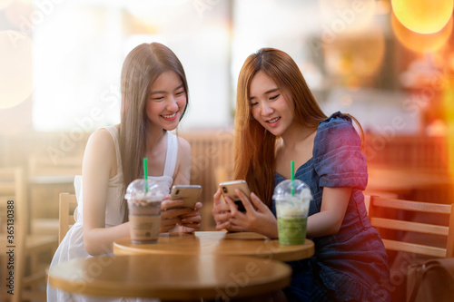 two happy beautiful young asian women sitting at table chatting talking playing with cellphone in coffee shop or tea house, shot through window glass.