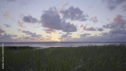 Forward and upward moving wide shot over long, green grass revealing the beach and ocean at Schiermonnikoog, the Netherlands. Sun setting against a blue, partly cloudy sky, photo