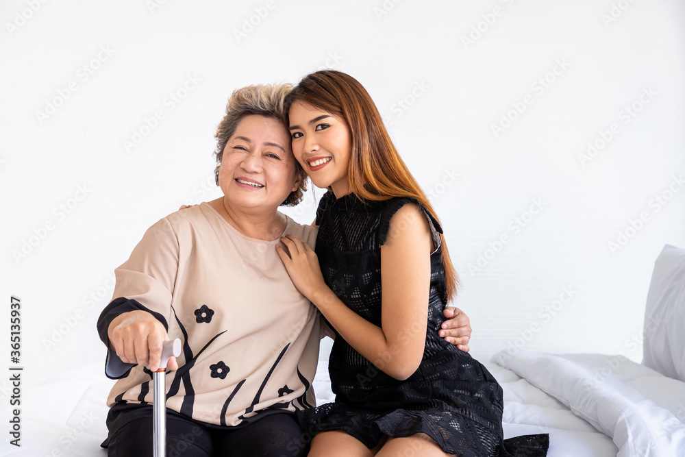 Fototapeta premium Happy senior elderly Asian woman female mother holding cane with daughter sitting on bed in bedroom, looking at camera