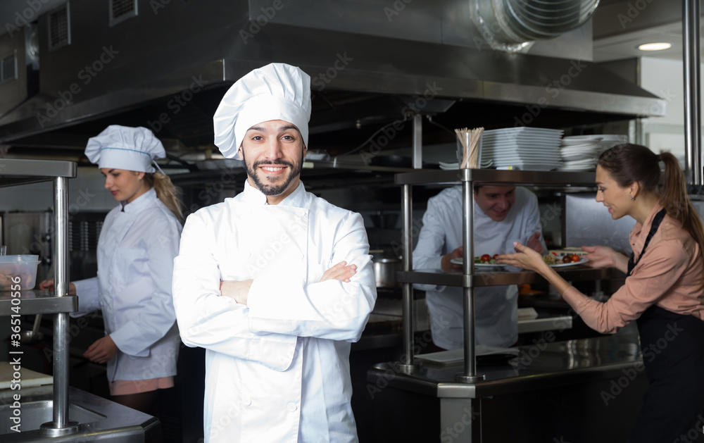 Portrait of satisfied smiling chef on restaurant kitchen with busy professional staff