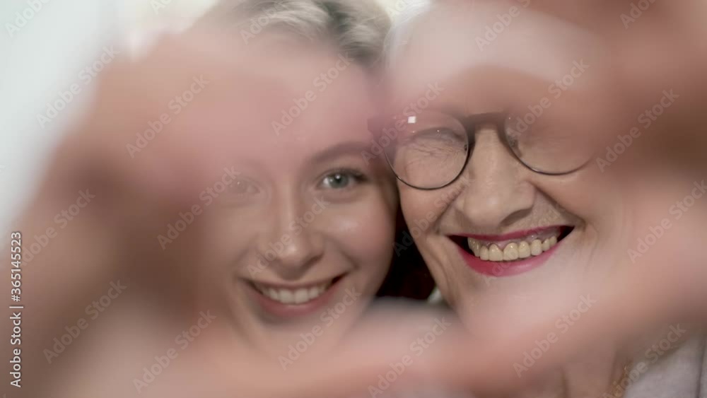 Smiling elderly grandmother and granddaughter make a heart-shaped hand gesture. Look at the camera connecting laughter showing the concept of family love, close-up portrait. Smiling elderly grandmother and granddaughter make a heart-shaped hand gesture. Look at the camera connecting laughter showing the concept of family love, close-up portrait.