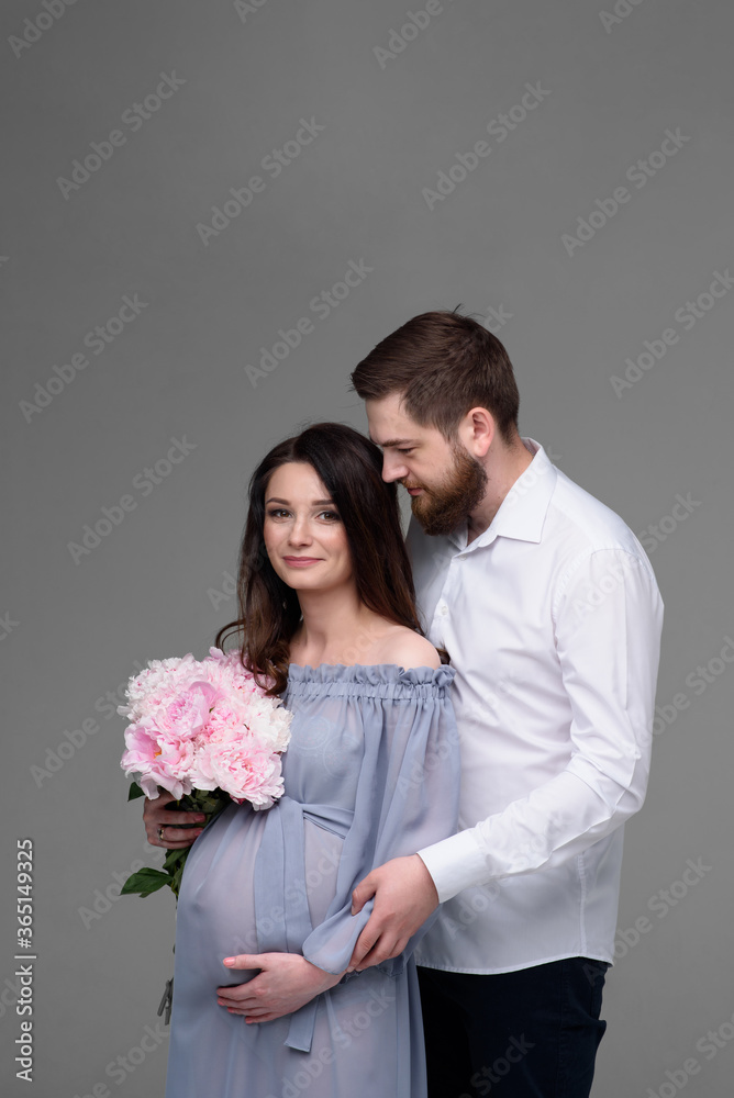 A pregnant woman and her husband are hugging on a gray background in the studio. A pregnant woman and her husband are hugging on a gray background in the studio.