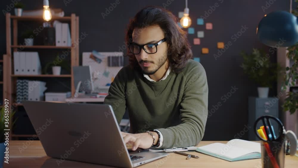 Slow motion of good-looking Arab guy in glasses working with laptop and taking notes in office at night. Career, employment and businesspeople concept. Slow motion of good-looking Arab guy in glasses working with laptop and taking notes in office at night. Career, employment and businesspeople concept.